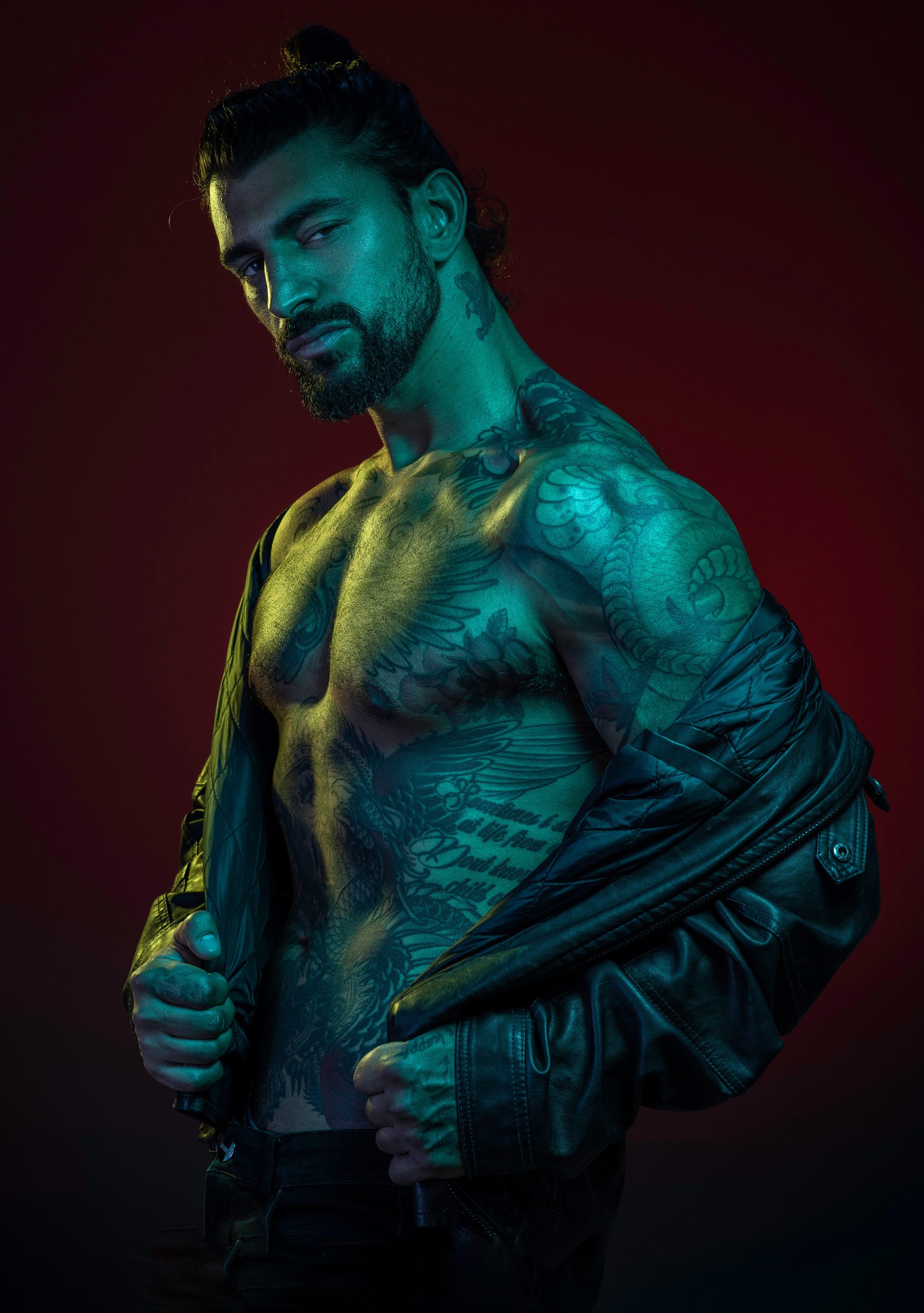 A male with muscular features covered with tattoos posing for a picture with dark red background and blue green gel color